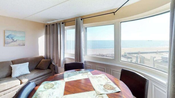 Een kamer met een ronde tafel, een bank en grote ramen met uitzicht op zee.
