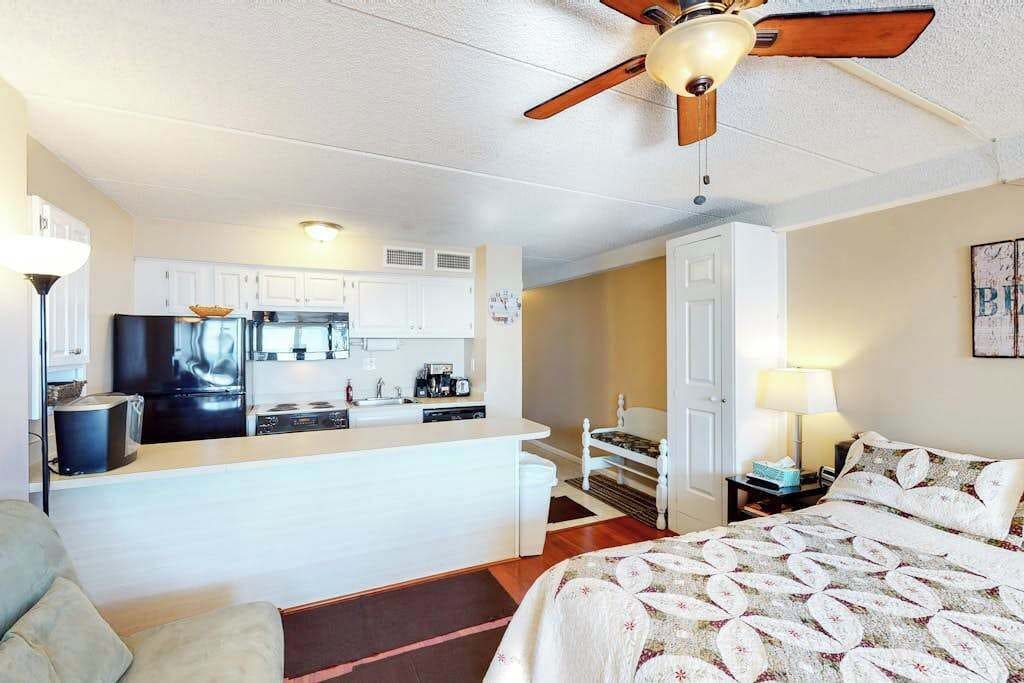 A compact hotel room with a kitchenette, a bed with a patterned bedspread, a ceiling fan, and a nightstand with a lamp.