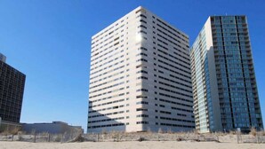 Een hoog, rechthoekig gebouw met een raster van ramen, omgeven door een zandig gebied met schaarse begroeiing.
