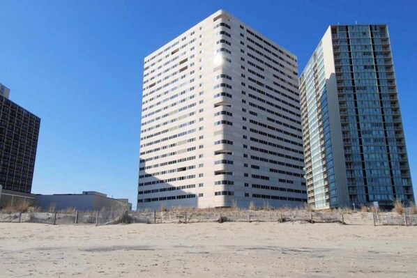 A tall, rectangular building with a grid of windows, surrounded by a sandy area with sparse vegetation.