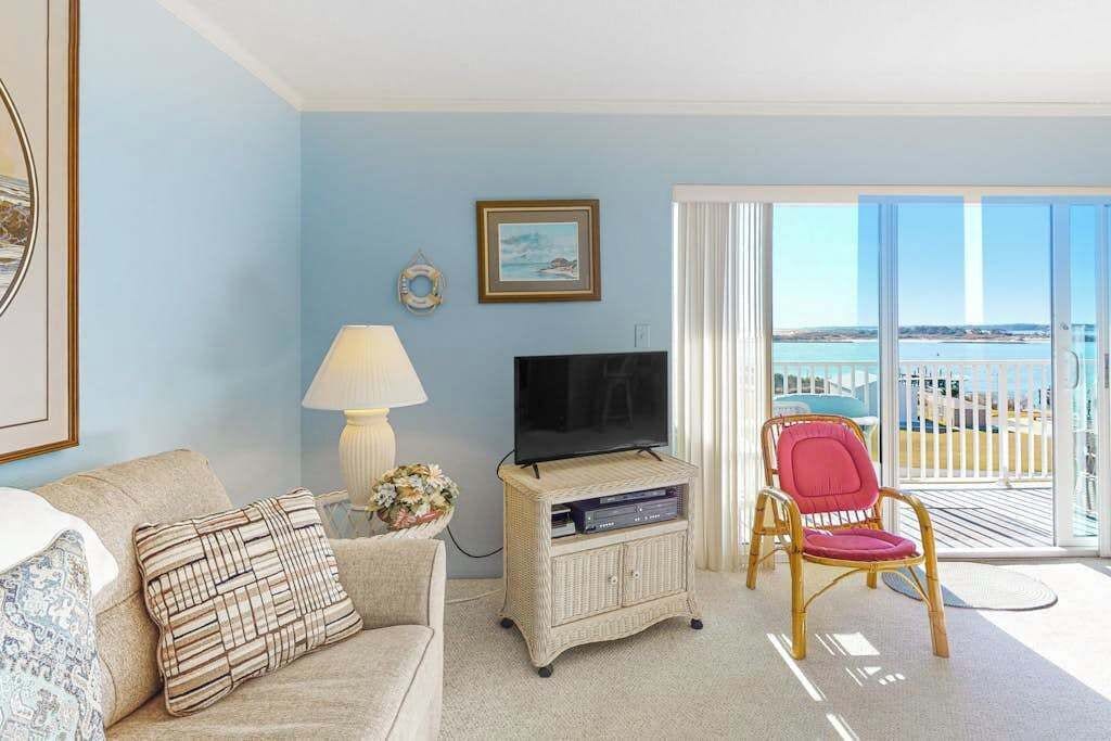 A living room with a television, a sofa, a chair, and a view of the sea through large windows.