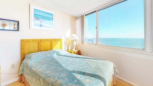 Un dormitorio con cama, ventana con vista al mar, un cuadro enmarcado en la pared y una mesita de noche con lámpara.