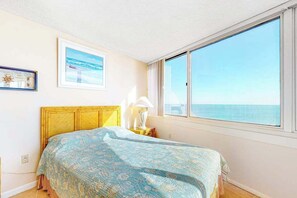Ein Schlafzimmer mit Bett, einem Fenster mit Meerblick, einem gerahmten Bild an der Wand und einem Nachttisch mit Lampe.