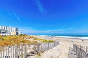 Ein Strand mit einer Holzumzäunung, einem Sandweg und einem mehrstöckigen Gebäude im Hintergrund.