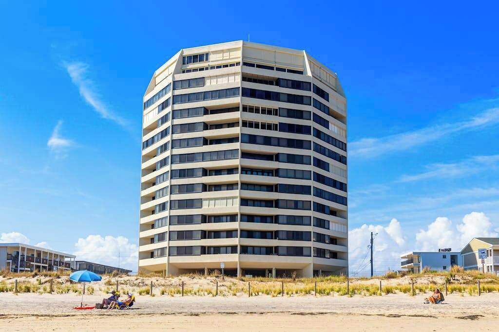 A multi-story hotel with a curved facade, located near a beach with a few people and a blue umbrella.