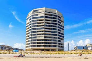 Ein mehrstöckiges Hotel mit geschwungener Fassade in Strandnähe, mit wenigen Personen und einem blauen Sonnenschirm.
