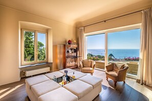 Ein geräumiger Raum mit großem Fenster, einer Couch, einem Sessel und einem kleinen Tisch mit Flaschen.