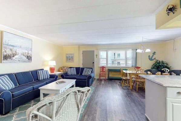 A living room with a sofa, coffee table, wicker chair, dining area with chairs and table, and a kitchen counter. - Ann Elizabeth 204 (Ocean City)