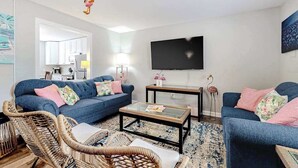 Sala de estar con sofá, mesa de centro, sillas de mimbre, televisor de pantalla plana y cocina al fondo.