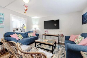 A living room with a sofa, coffee table, wicker chairs, a flat-screen TV, and a kitchen in the background. - Flamingo House (Ocean City)
