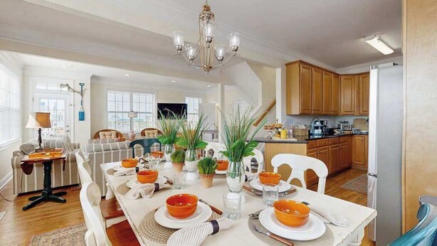 Comedor con una larga mesa dispuesta para seis personas, rodeada de sillas. Encima de la mesa hay una araña, y en el fondo se aprecia una cocina y armarios de madera.