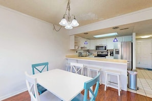 A compact kitchen with white cabinetry, a central island, and a dining area with a white table and blue chairs. - Top-floor, corner condo steps to beach with balcony & ocean views (Ocean City)
