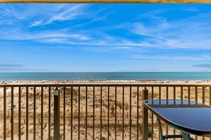 Blick auf einen Strand vom Balkon aus, mit Tisch und Stühlen, einem Zaun und wolkenlosem Himmel.
