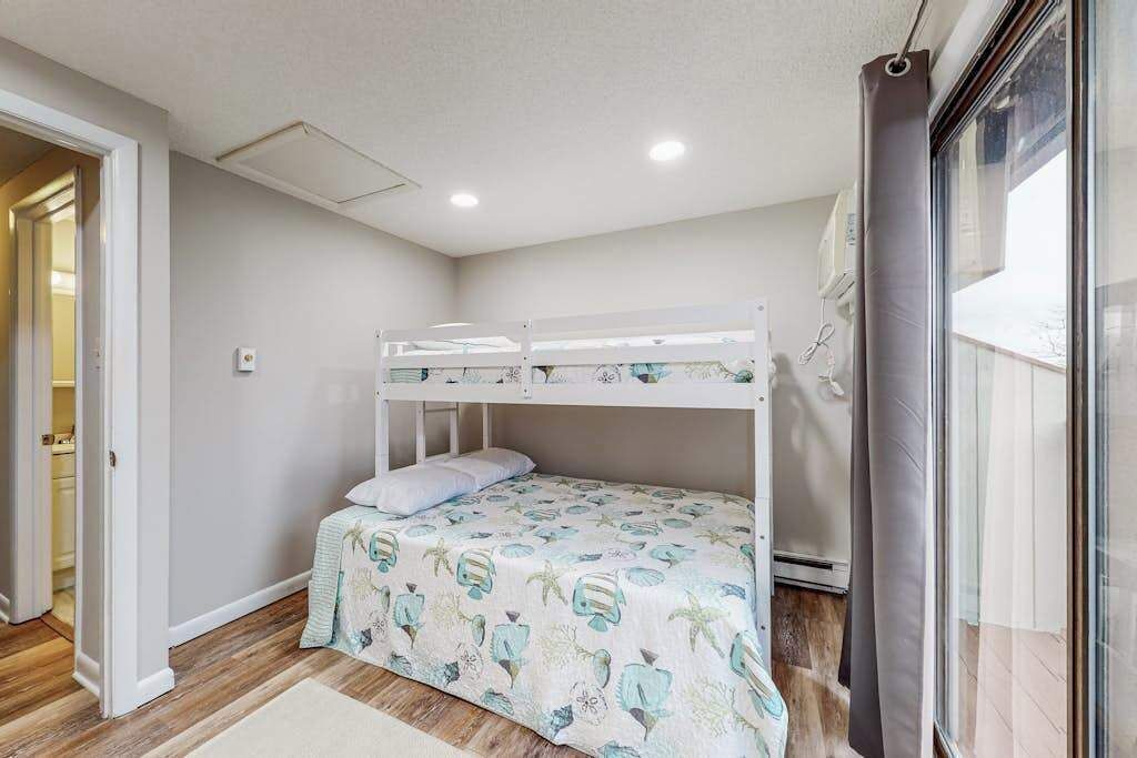Uma cama de beliche com colcha florida, piso de madeira, porta branca e janela com cortinas.