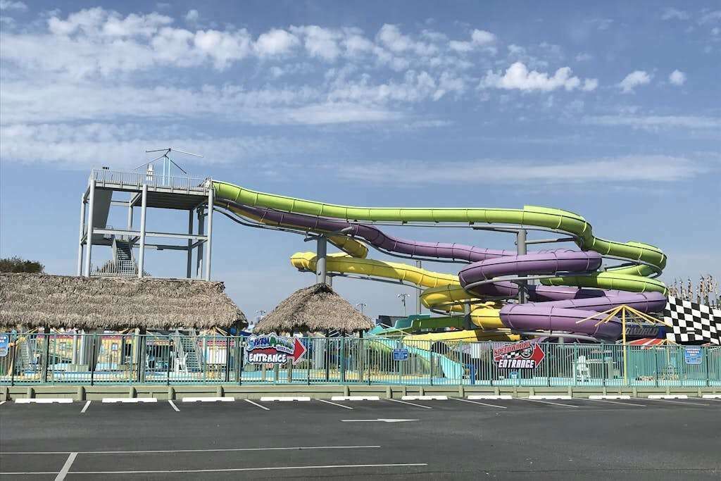 Un parque acuático con un tobogán de agua grande y de varios colores, una estructura con techo de paja y un estacionamiento cercado.