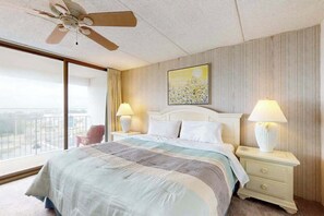 A hotel room with a bed, two bedside lamps, a ceiling fan, a large window, and a painting on the wall. - Bay-view 5th-floor condo with balcony, AC, indoor pool & gym - walk to beach (Ocean City)