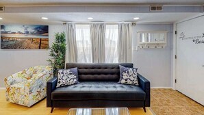 Un salon avec un canapé noir, un pouf à motifs, un grand tableau et un mur orné d’un panneau décoratif.