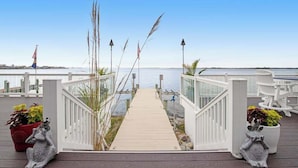 A wooden deck leading to a waterfront with a railing, potted plants, and flags. - A Bayfront Row Seat (Ocean City)