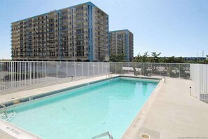 A swimming pool with a white fence, a pool ladder, and a building with balconies in the background. - Excalibur 905 (Ocean City)