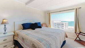 A bedroom with a bed, a nightstand, a lamp, a window with curtains, and a view of the sea. - Excalibur 905 (Ocean City)