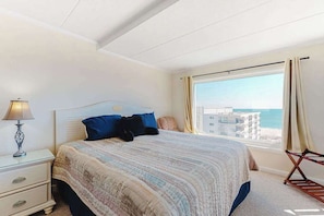 A bedroom with a bed, a nightstand, a lamp, a window with curtains, and a view of the sea. - Fantastic Condo Situated in Oceanfront Complex w/Shared Pool, Free WiFi, AC (Ocean City)