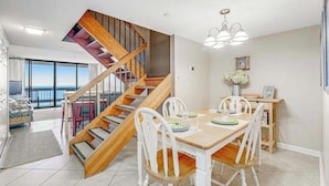 A dining area with a wooden staircase, white chairs, and a table set for four. - 22nd-Floor Oceanfront Gem with Three Balconies, W/D, AC, and Shared Heated Pool (Ocean City)