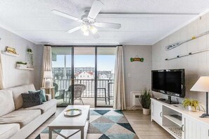 A modern living room with a sofa, coffee table, TV, and balcony with a city view. - Bright, sunny condo with ocean and bay views in walkable Midtown location (Ocean City)