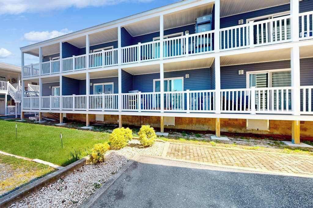 A multi-story building with balconies, a paved driveway, and a grassy area with small bushes.