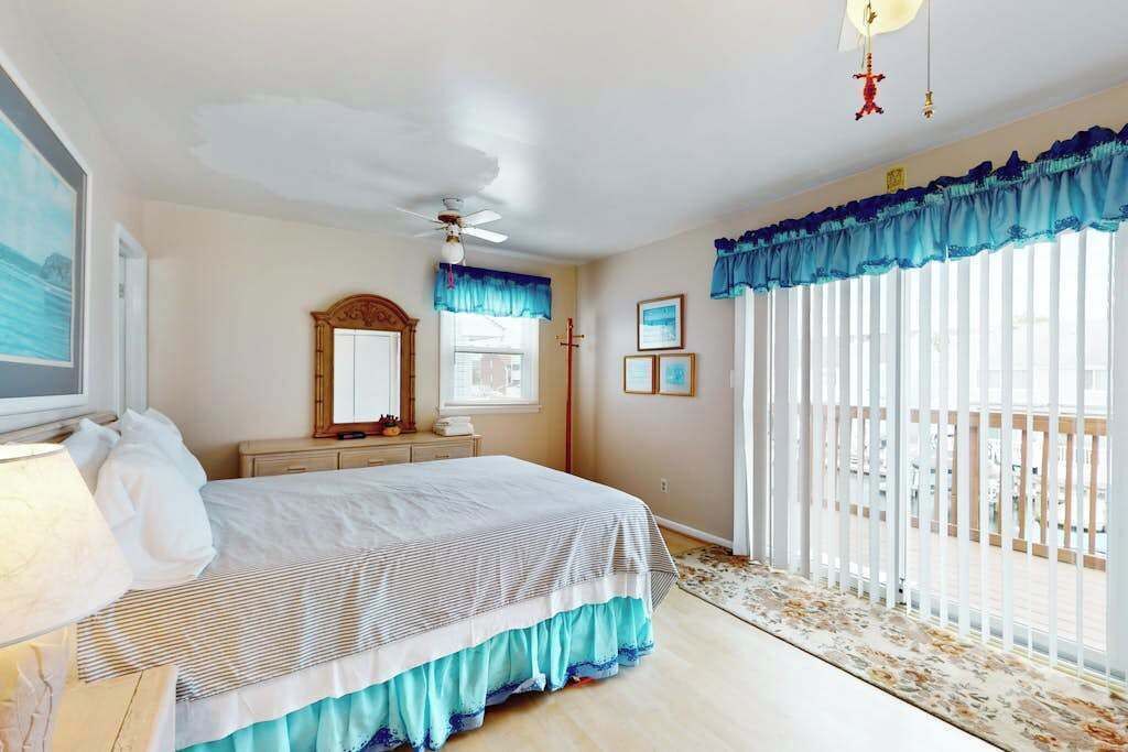 A bedroom with a bed, a ceiling fan, a dresser, a window with curtains, and a view of the outside.