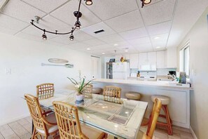 A dining area with a glass table and wicker chairs, a kitchen with white cabinets, and a ceiling with recessed lighting. - Waikiki 406 (Ocean City)