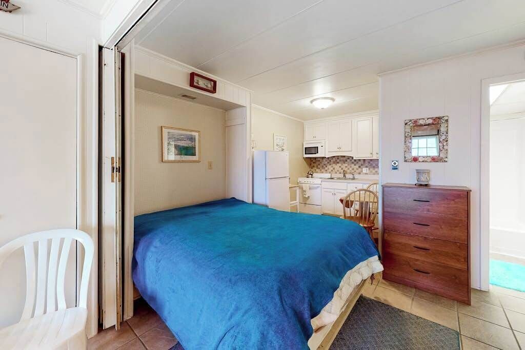A bedroom with a bed, a wooden dresser, a white chair, a kitchenette with a microwave, and a small window.