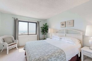 A bedroom with a bed, a chair, a nightstand, a lamp, a window, and a plant. - Oceanfront Condo with Balcony, WiFi, W/D & Shared Pool - Close to Boardwalk (Ocean City)