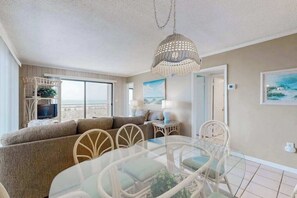 A living room with a sofa, a glass-top dining table, wicker chairs, a hanging light fixture, and a television. - Oceanfront Condo with Balcony, WiFi, W/D & Shared Pool - Close to Boardwalk (Ocean City)