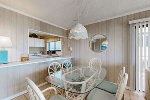 A dining area with a glass table and wicker chairs, a kitchen with white cabinets, and a hanging light fixture. - Oceanfront Condo with Balcony, WiFi, W/D & Shared Pool - Close to Boardwalk (Ocean City)