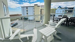 En balkong med vita plaststolar och ett bord, med utsikt över en parkeringsplats och byggnader, och därifrån ser man även stranden.