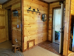 A wooden interior with a wooden bench, a small table, a wall-mounted mirror, and a door leading to another room.