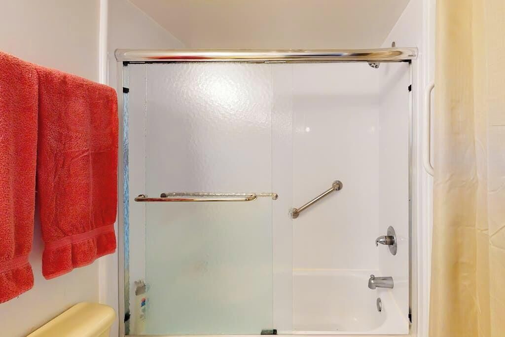 Eine Duschkabine mit Glas, eine Metallhandstütze, eine weiße Badewanne und ein rotes Handtuch an der Wand.