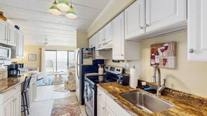 A kitchen with white cabinets, a stainless steel sink, a microwave, a refrigerator, and a dining area with a table and chairs. - West Winds 404 | Condo in Ocean City (Ocean City)