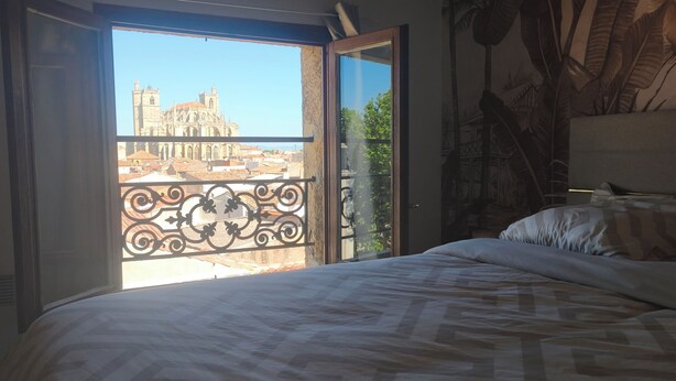 Une chambre à coucher avec un grand lit, un balcon doté d’une balustrade en fer forgé et une vue sur un bâtiment historique.