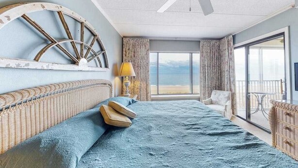 Habitación de hotel con cama, ventilador de techo, ventana grande con vistas y una silla de mimbre.