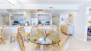 Una cocina moderna con armarios blancos, una isla central y un comedor con mesa de vidrio y sillas de mimbre.