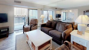 Ein Wohnzimmer mit Sofa, Sessel, Couchtisch, Fernseher und einem Balkon mit Blick auf Gebäude.