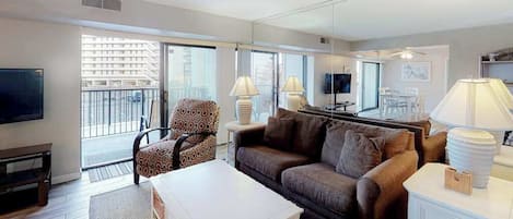 A living room with a sofa, armchair, coffee table, TV, and a balcony with a view of buildings.