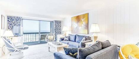 A living room with a sofa, armchairs, a coffee table, and a TV. There is a balcony with a view.