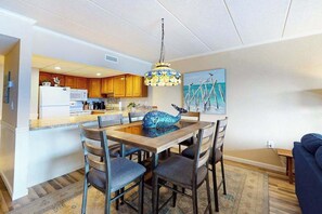 A modern kitchen with wooden cabinets, a dining area with a glass table, and a painting on the wall. - Coastal Condo w/Side Ocean Views, Balcony, Shared Pool, Private W/D, Free WiFi (Ocean City)