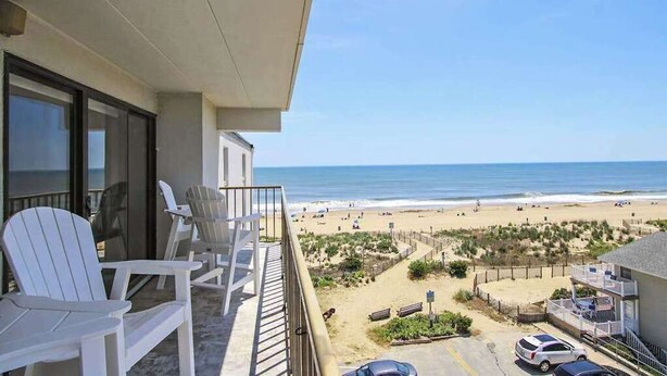 Un balcó amb caires blanques que dona a una platja, amb una tanca, una zona d'aparcament i un edifici amb balcó.