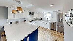 Une cuisine moderne avec des armoires blanches, un grand îlot central, des appareils en acier inoxydable et des luminaires suspendus.