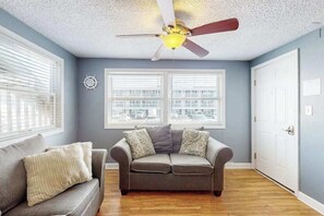 Ein Zimmer mit grauem Sofa, einem gemusterten Sessel, einem Deckenventilator und zwei Fenstern mit Jalousien.