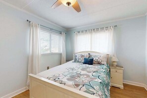 Ein Schlafzimmer mit Bett, Nachttisch, Deckenventilator und zwei mit Vorhängen versehenen Fenstern.