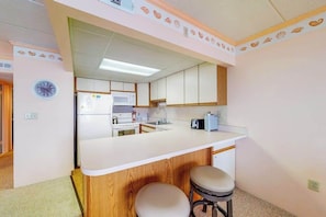 A kitchen with a white countertop, wooden cabinets, a microwave, and a sink. There are two stools and a clock on the wall. - Bay view condo with WiFi, balcony, boardwalk, pool, tennis, & on-site dining (Ocean City)